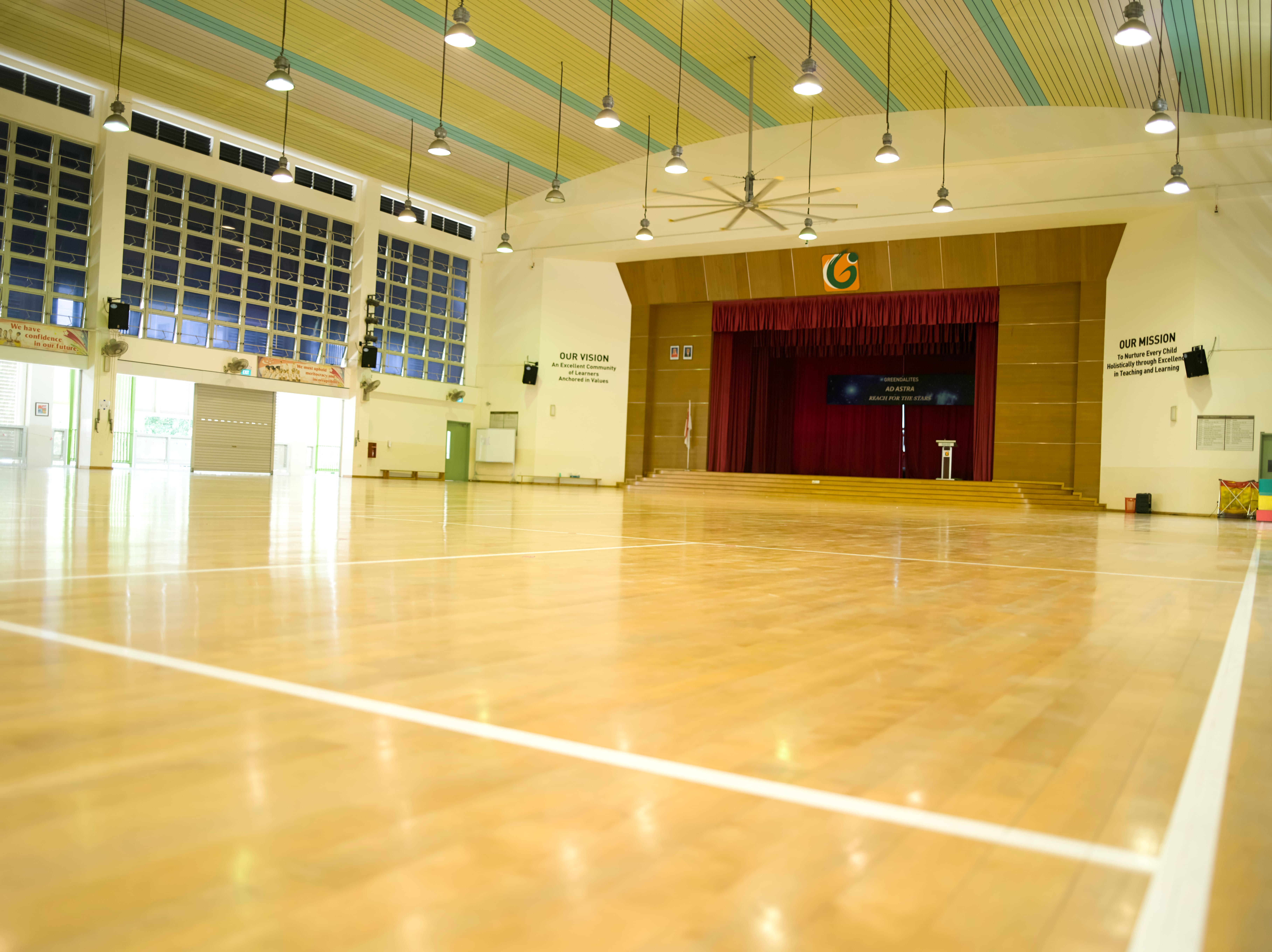 Spacious school hall with a stage, wooden flooring and high ceiling.